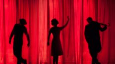 Tres siluetas de personas detrás de una cortina roja. Teatro para la inclusión de personas sordas