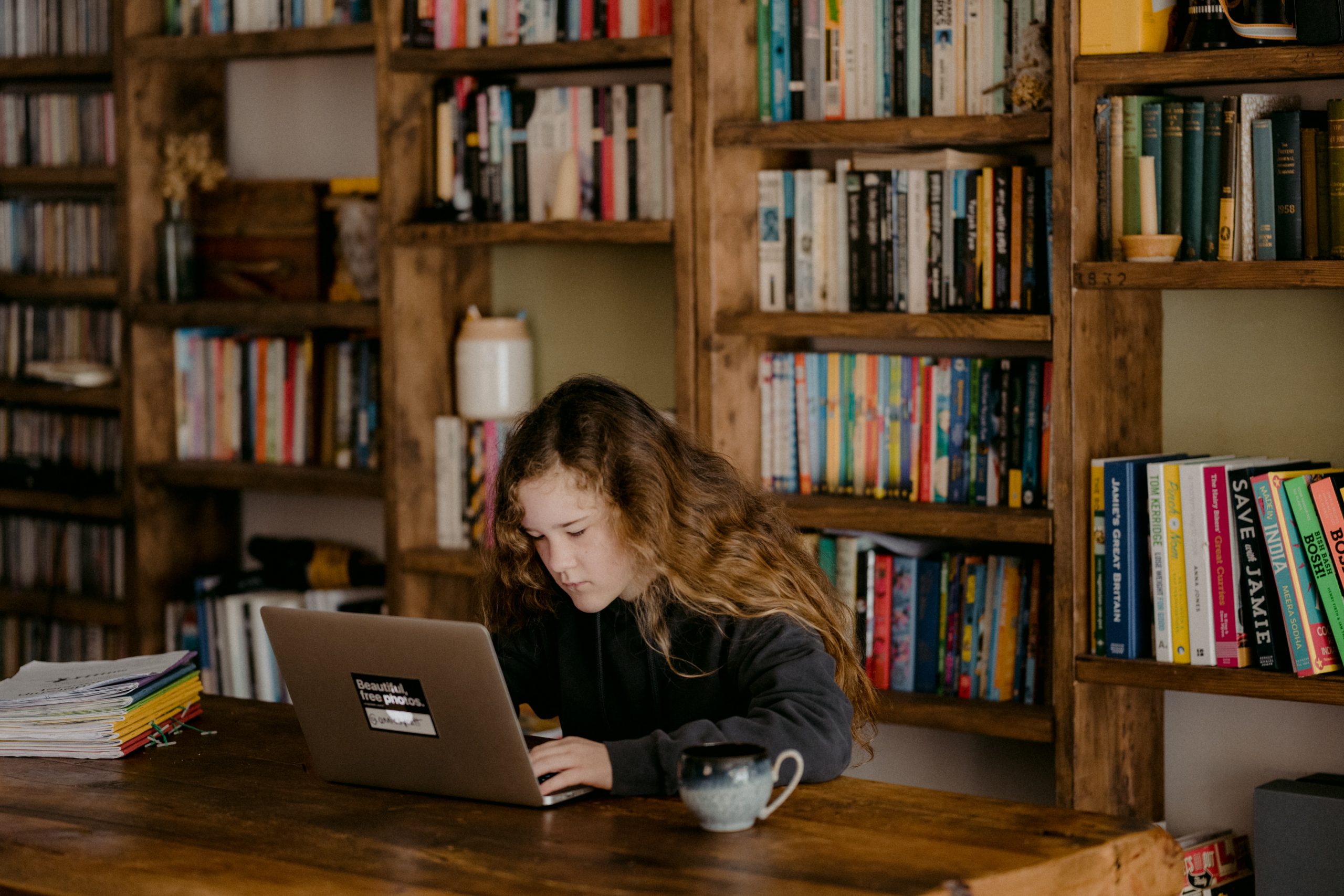 Niña con un computador, sentada en un escritorio con una biblioteca de fondo. Educación desde casa